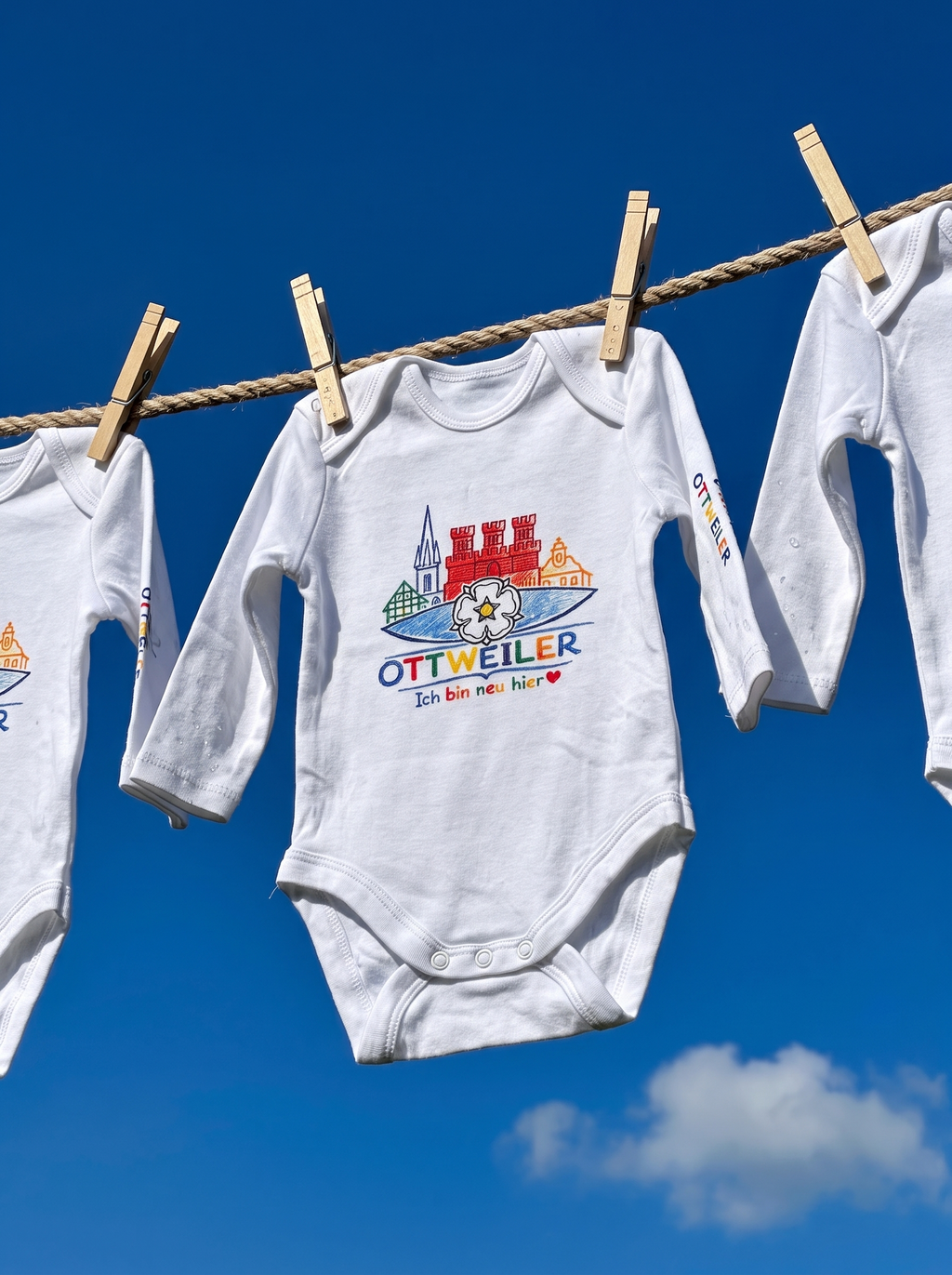Drei weiße Ottweiler Baby-Bodys mit Stadtmotiv an einer Wäscheleine vor blauem Himmel